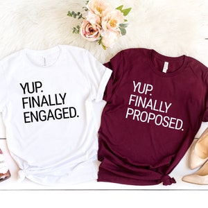 May include: Two white t-shirts with black text. The left shirt says "YUP. FINALLY ENGAGED." The right shirt says "YUP. FINALLY PROPOSED."