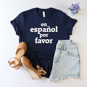 May include: A navy blue t-shirt with the text "en espa&ntilde;ol por favor" in white. The shirt is paired with denim shorts and brown sandals.