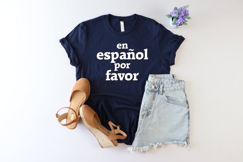 May include: A navy blue t-shirt with the text "en espa&ntilde;ol por favor" in white. The shirt is paired with denim shorts and brown sandals.