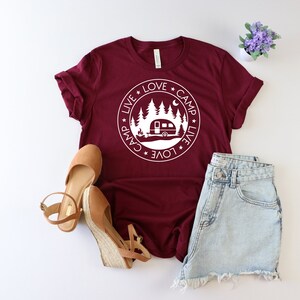 May include: A maroon t-shirt with a white circle graphic featuring a camper, trees, and the text "Live * Love * Camp * Live * Love * Camp".