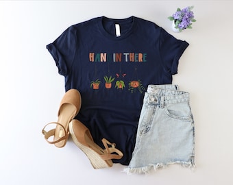 May include: A navy blue t-shirt with the text "HANG IN THERE" in colorful letters. The text is surrounded by a graphic of hanging plants in pots.