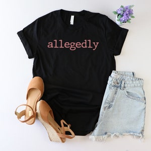 Op de afbeelding: Zwart T-shirt met het woord "allegedly" in roze op de voorkant. Het shirt wordt gecombineerd met een spijkerbroekje en bruine wedges.