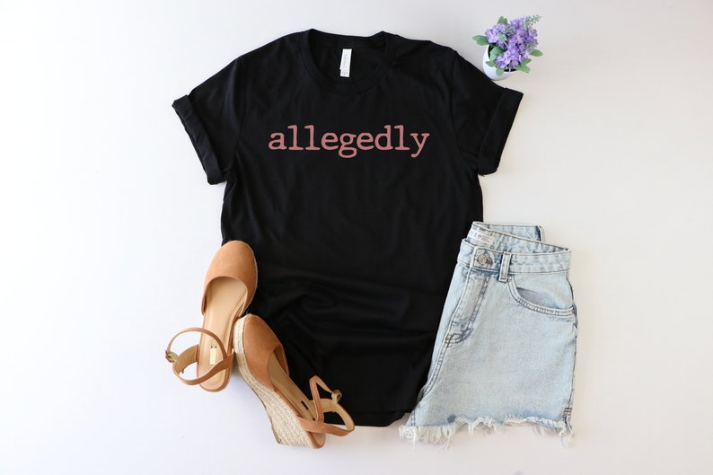 Op de afbeelding: Zwart T-shirt met het woord "allegedly" in roze op de voorkant. Het shirt wordt gecombineerd met een spijkerbroekje en bruine wedges.