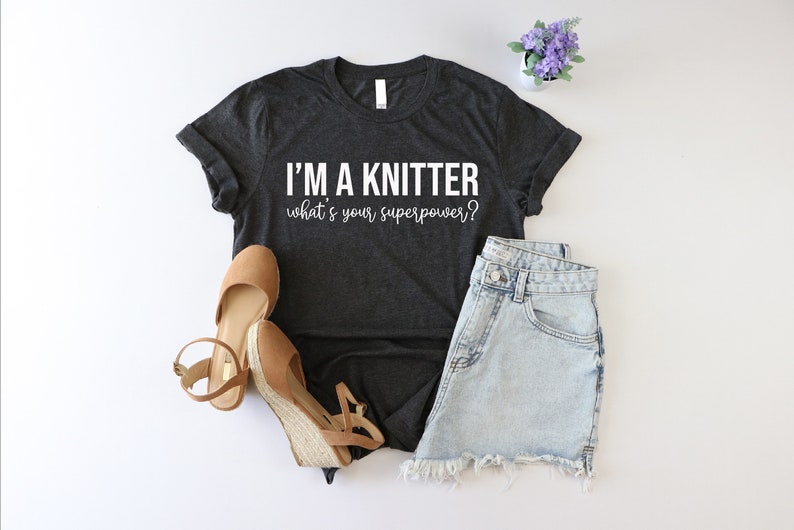 May include: A dark gray t-shirt with the text "I'm a knitter what's your superpower?" in white. The shirt is paired with denim shorts and brown wedges.