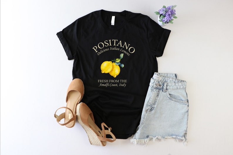 Może przedstawiać: Czarny T-shirt z białym nadrukiem, na kt&oacute;rym widnieje napis "Positano Delicious Italian Lemons" i "Fresh from the Amalfi Coast, Italy". Nadruk zawiera r&oacute;wnież dwa ż&oacute;łte cytryny z zielonymi liśćmi.