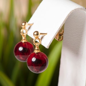 Peut inclure: Boucles d'oreilles pendantes dorées avec des perles rondes rouge foncé, semblables à du verre. Les boucles d'oreilles sont présentées sur un support blanc sur un fond vert et jaune flou. Fermeture à tige et boule.