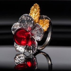 May include: A silver ring featuring a red gemstone and gold leaf accents, shaped like a flower.