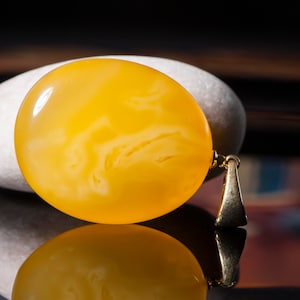 Op de afbeelding: Een gele amber hanger met een swirl-patroon en een gouden balein. De hanger ligt op een witte steen.