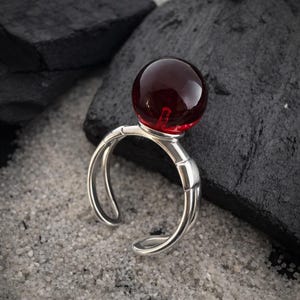 May include: A silver-toned ring featuring a smooth, spherical dark red glass bead set atop a split-shank band. The metal band has a polished finish and an open design, showcasing a reflective surface that contrasts with the deep red orb.