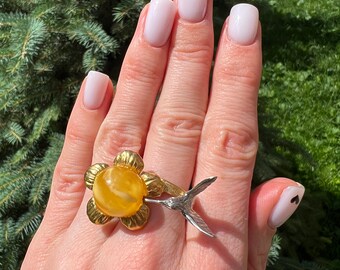 Könnte beinhalten: Ein goldener Ring mit einem gelben Bernstein in Form einer Blume und einem silbernen Kolibri-Schwanz.
