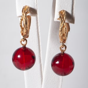 May include: A pair of gold-toned earrings featuring a textured design at the top and a round, deep red glass bead. The earrings are displayed on a white stand against a white background. The beads are approximately 1.5 cm in diameter.