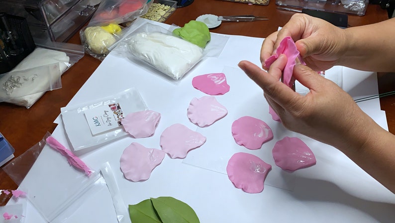May include: A close-up of a person's hands shaping pink polymer clay petals. The petals are arranged on a white surface, along with a green leaf and a bag of white polymer clay. The bag has the text "Joyful Clay" and "PMC" printed on it.