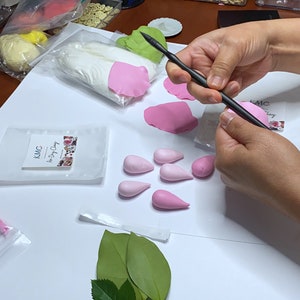 May include: A person is using a tool to shape pink polymer clay into flower petals. There are several other pink clay petals and a bag of white polymer clay on the table. The table is covered in white paper.