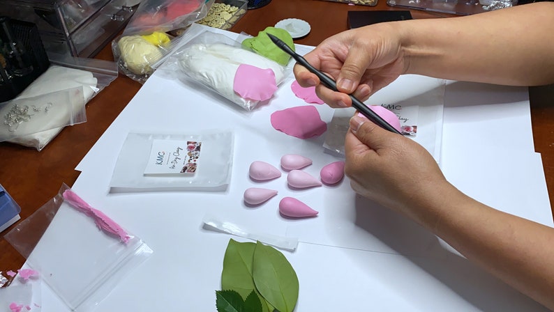 May include: A person is using a tool to shape pink polymer clay into flower petals. There are several other pink clay petals and a bag of white polymer clay on the table. The table is covered in white paper.