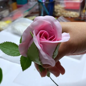 May include: A pink rose made of polymer clay with green leaves attached to a wire stem. The rose is held in a hand.