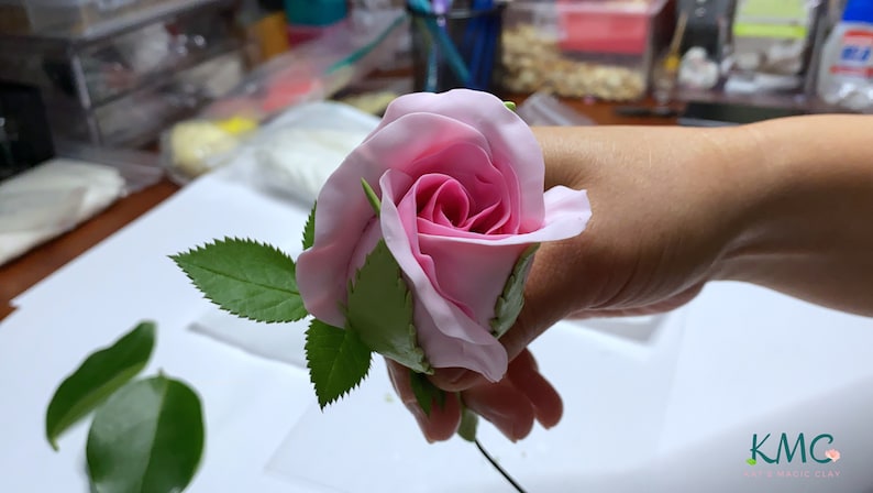 May include: A pink rose made of polymer clay with green leaves attached to a wire stem. The rose is held in a hand.