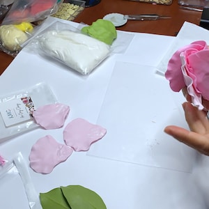 May include: A pink sugar flower with a white center is being held by a person's hand. The flower is made of fondant and is being assembled on a white surface. There are other pink sugar flower petals and green leaves on the surface.