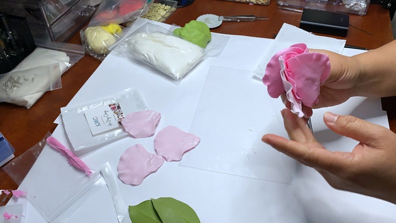 May include: A pink sugar flower with a white center is being held by a person's hand. The flower is made of fondant and is being assembled on a white surface. There are other pink sugar flower petals and green leaves on the surface.