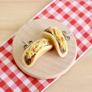 May include: A pair of taco earrings made of polymer clay. The earrings are shaped like tacos with fillings of ground beef, lettuce, cheese, and red and yellow sprinkles. The earrings are on a wooden surface with a red and white checkered background.