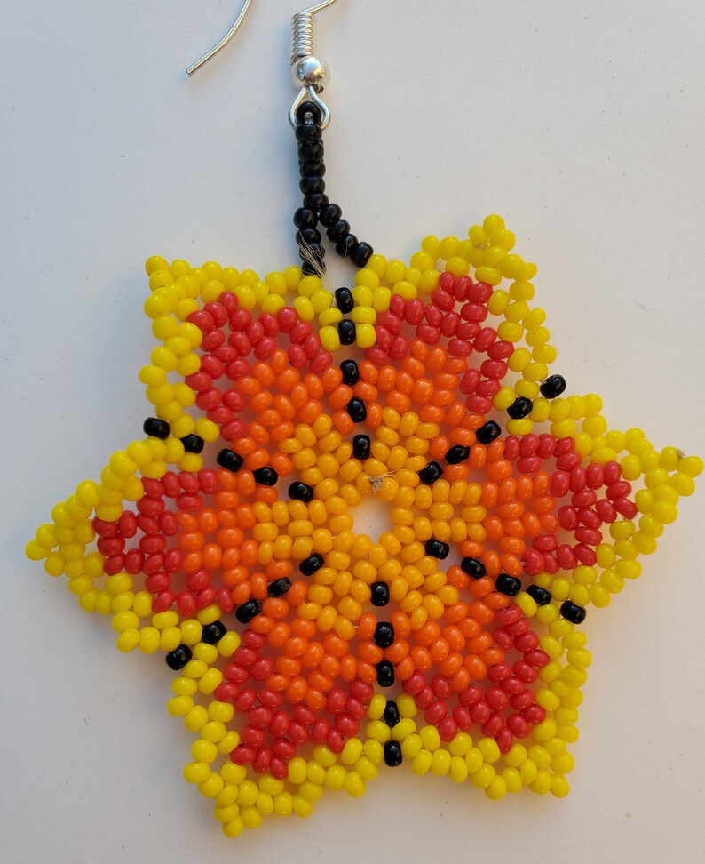 Puede incluir: Un pendiente de estrella con cuentas de color amarillo, naranja y rojo degradado. El centro de la estrella tiene un patr&oacute;n de cuentas negras.
