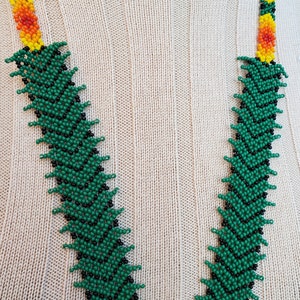 Puede incluir: Un collar de cuentas verde, negro y amarillo con un patr&oacute;n en zigzag. El collar est&aacute; sobre un fondo de tejido de punto beige.