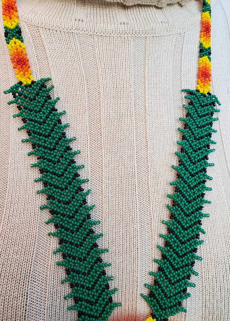 Puede incluir: Un collar de cuentas verde, negro y amarillo con un patr&oacute;n en zigzag. El collar est&aacute; sobre un fondo de tejido de punto beige.