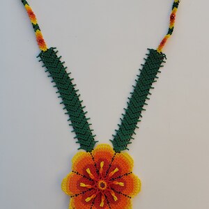 Puede incluir: Un collar de cuentas con un gran colgante de flor naranja. El collar est&aacute; hecho de cuentas verdes, amarillas y naranjas. El colgante de flor tiene una franja de cuentas.
