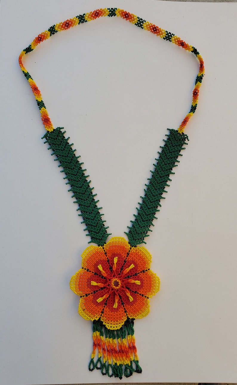 Puede incluir: Un collar de cuentas con un gran colgante de flor naranja. El collar est&aacute; hecho de cuentas verdes, amarillas y naranjas. El colgante de flor tiene una franja de cuentas.