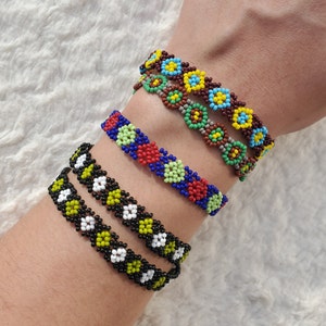 Puede incluir: Cuatro pulseras de cuentas con coloridos patrones de flores. Las pulseras están apiladas en la muñeca de una persona. Las pulseras están hechas con cuentas negras, blancas, rojas, azules, verdes y amarillas.