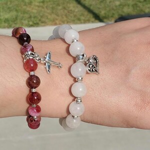 Puede incluir: Dos pulseras de cuentas en una muñeca. La pulsera de la izquierda está hecha de cuentas rojas y rosadas con un dije de avión plateado. La pulsera de la derecha está hecha de cuentas blancas con un dije de corazón plateado.