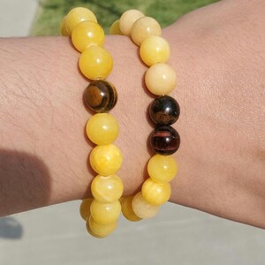 Puede incluir: Dos pulseras de cuentas en una muñeca. Una pulsera está hecha de cuentas amarillas y la otra de cuentas blancas. Ambas pulseras tienen una cuenta marrón oscura en el centro.