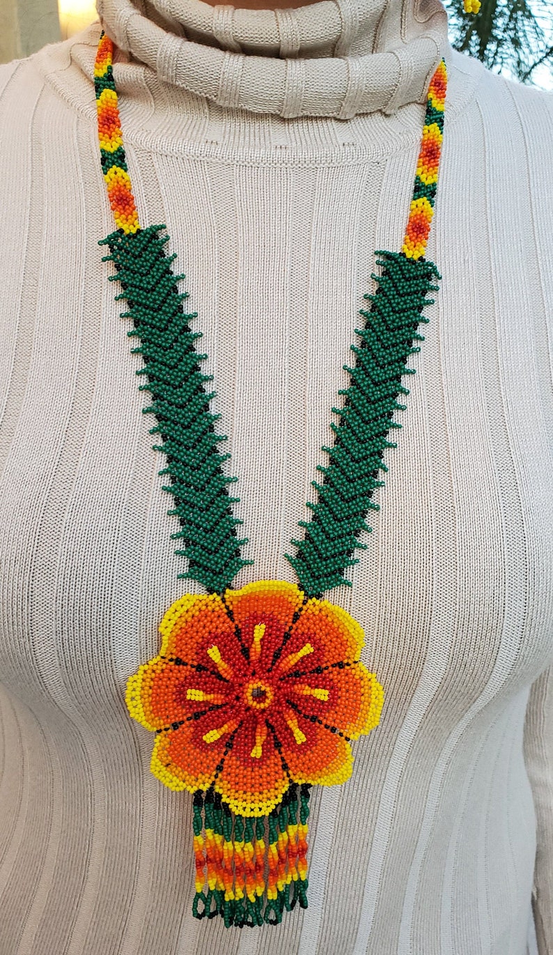 Puede incluir: Un collar de cuentas con un gran colgante de flor naranja, amarilla y verde. El collar est&aacute; hecho de cuentas verdes y tiene una franja de cuentas naranja, amarillas y verdes.