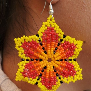 Puede incluir: Un par de pendientes en forma de estrella hechos con cuentas de colores. Los pendientes son amarillos, naranjas, rojos y negros. Los pendientes cuelgan de ganchos plateados.