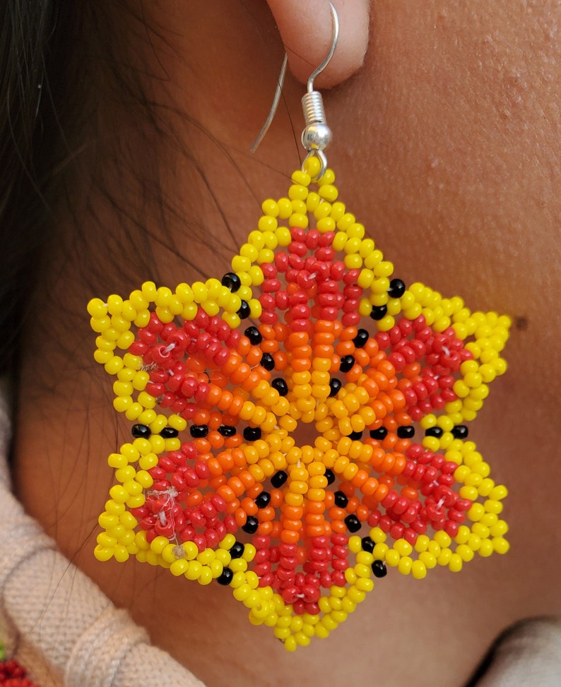 Puede incluir: Un par de pendientes en forma de estrella hechos con cuentas de colores. Los pendientes son amarillos, naranjas, rojos y negros. Los pendientes cuelgan de ganchos plateados.