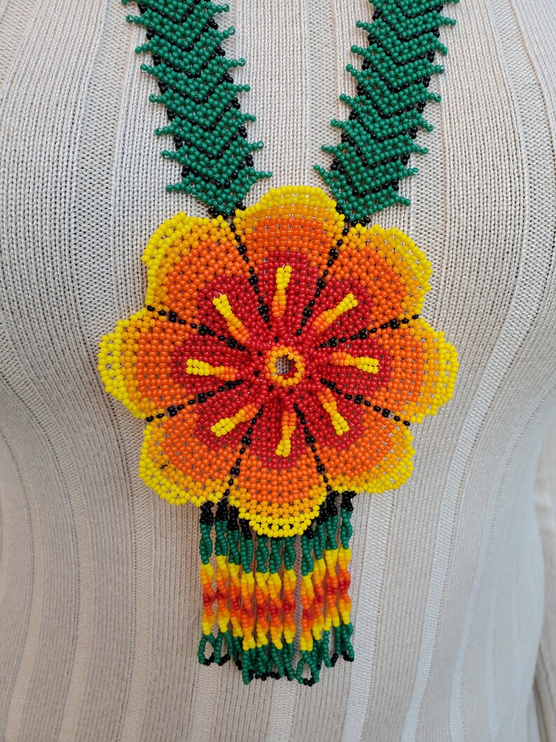 Puede incluir: Un collar de cuentas con un gran colgante de flor. La flor est&aacute; hecha de cuentas de color naranja, amarillo, rojo y verde. El collar tiene una franja de cuentas verdes, naranjas, amarillas y rojas.