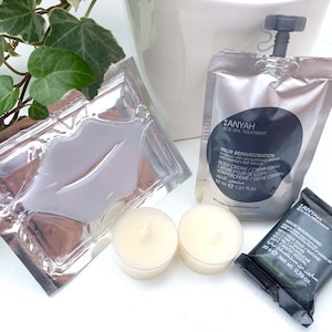 May include: A set of spa products in individual packaging. The set includes a lip mask, a body cream, a bar of soap, and two small candles. The products are all in silver and black packaging with the brand name "ANYAH" printed on them.