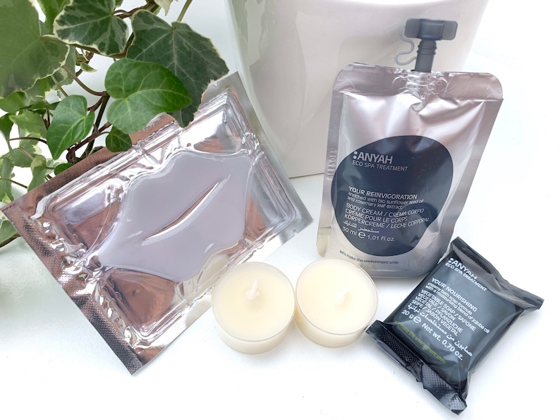 May include: A set of spa products in individual packaging. The set includes a lip mask, a body cream, a bar of soap, and two small candles. The products are all in silver and black packaging with the brand name "ANYAH" printed on them.