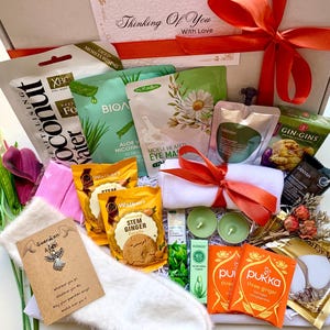 May include: A wellness gift box containing face masks, tea, ginger biscuits, candles, and a small towel tied with an orange ribbon. The box is decorated with a large orange bow and a card that says "Thinking Of You With Love."
