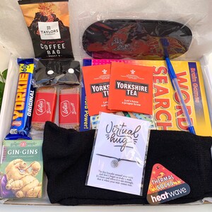 May include: A gift box filled with various items, including a black eye mask, a black pair of socks, a small blue lip balm, a pack of Yorkshire Tea, a chocolate bar, a pack of biscuits, a small book of word search puzzles, a pack of cards, a small bag of coffee, a bracelet with a silver charm, and a card with the text "Virtual hug".