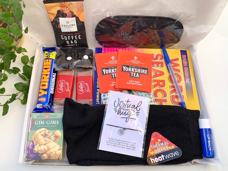 May include: A gift box filled with various items, including a black eye mask, a black pair of socks, a small blue lip balm, a pack of Yorkshire Tea, a chocolate bar, a pack of biscuits, a small book of word search puzzles, a pack of cards, a small bag of coffee, a bracelet with a silver charm, and a card with the text "Virtual hug".