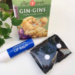 May include: A blue lip balm tube with the words "ORIGINAL LIP BALM" printed on it.  The lip balm is next to a black cloth face mask with two white buttons.  A box of "GIN-GINS CHEWY GINGER CANDY" is in the background.