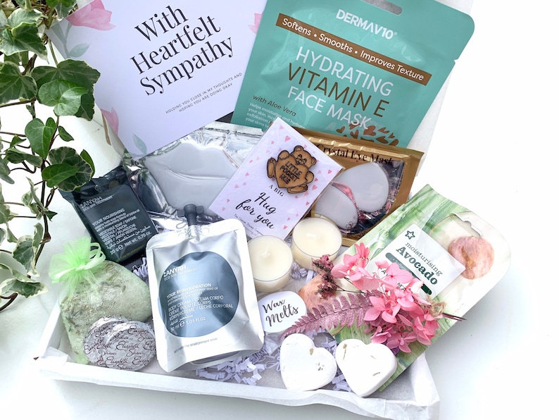 May include: A white gift box filled with self-care items, including a face mask, bath bombs, candles, and a wooden heart with the text "A big hug for you". The box also includes a card with the text "With Heartfelt Sympathy".