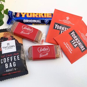 May include: A flat lay of various food and beverage items, including a chocolate bar labeled "Yorkie Original", two red and white packages of "Taylors of Harrogate Yorkshire Tea", a black coffee bag labeled "Hot Lava Java Coffee Bag - Roast 6", and two red and white packages of Lotus Biscoff cookies.