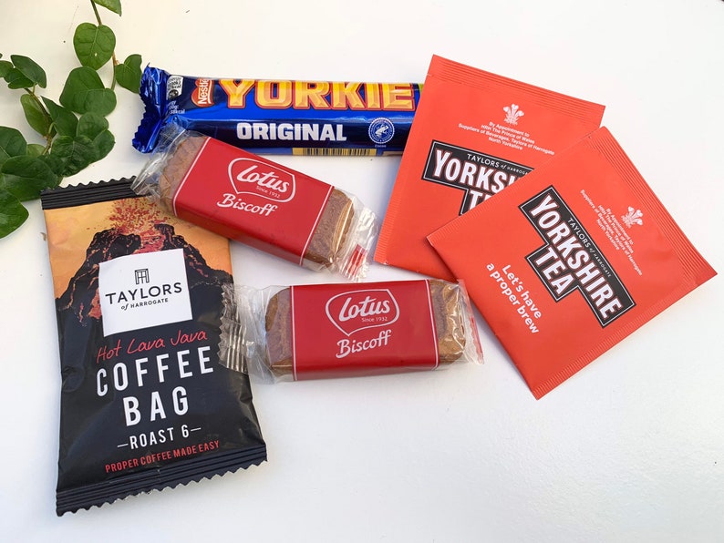 May include: A flat lay of various food and beverage items, including a chocolate bar labeled "Yorkie Original", two red and white packages of "Taylors of Harrogate Yorkshire Tea", a black coffee bag labeled "Hot Lava Java Coffee Bag - Roast 6", and two red and white packages of Lotus Biscoff cookies.