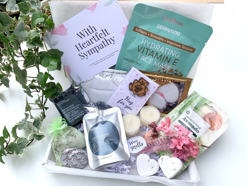May include: A white gift box filled with self-care items, including a face mask, bath bomb, eye mask, candles, and a wooden sign that says "A big hug for you". The box also includes a card that says "With Heartfelt Sympathy".