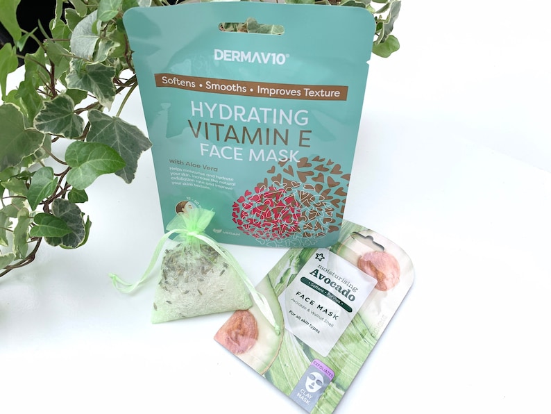May include: Two face masks, one with a green and white design and the text "Moisturizing Avocado Face Mask" and the other with a blue and gold design and the text "Hydrating Vitamin E Face Mask with Aloe Vera".