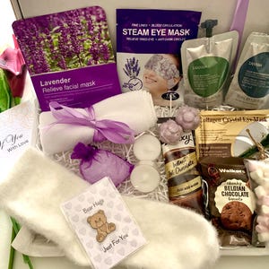 May include: A gift box containing self-care products. It features a lavender facial mask, steam eye mask, eye gels, a towel with a purple ribbon, candles, instant hot chocolate, Belgian chocolate biscuits, and a card that says "Bear Hugs Just For You."