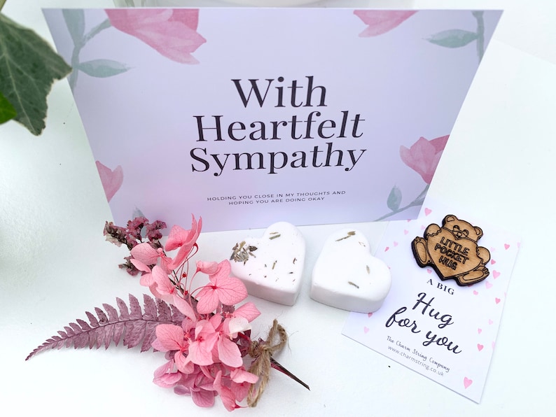 May include: A sympathy gift set with a card that reads "With Heartfelt Sympathy", two white heart-shaped bath bombs, a small wooden bear charm with the text "Little Pocket Hug", and a card that reads "A Big Hug for You".