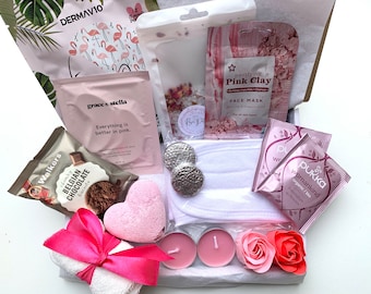 May include: A pink and white gift box filled with various self-care items, including a face mask, bath bomb, tea bags, and candles. The box also includes a pink towel, chocolate, and a pink heart-shaped bath bomb.