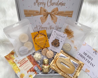 May include: A Christmas gift box filled with various items. Includes tea light candles, face mask, bath salts, tea, cookies, a gold bear, and lip masks. The box has a bow and the text "Merry Christmas & A Happy New Year".